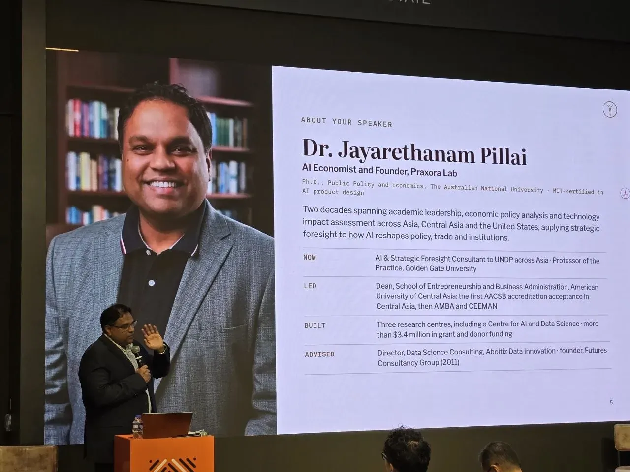 Dr. Jayarethanam Pillai speaking at the lectern in front of a screen showing his photograph and speaker profile.
