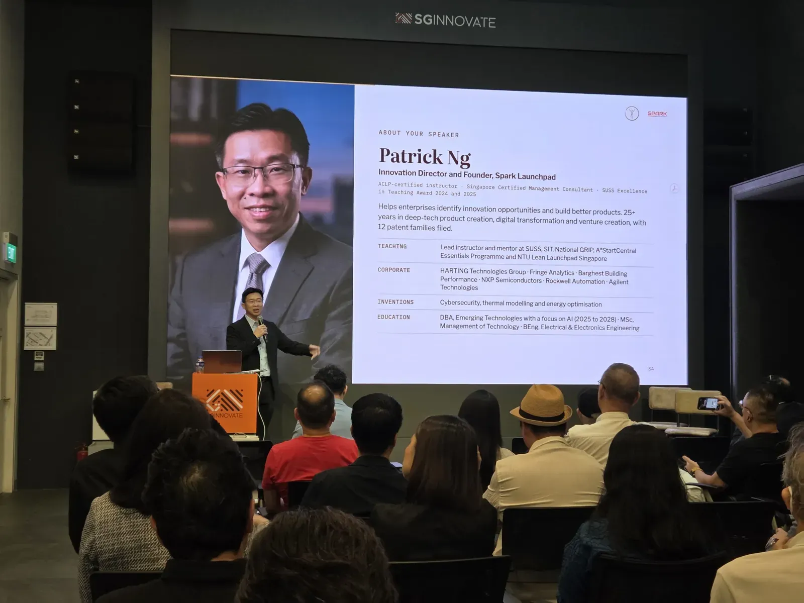 Patrick Ng at the lectern beneath a large screen showing his speaker profile, with the seated audience in the foreground.