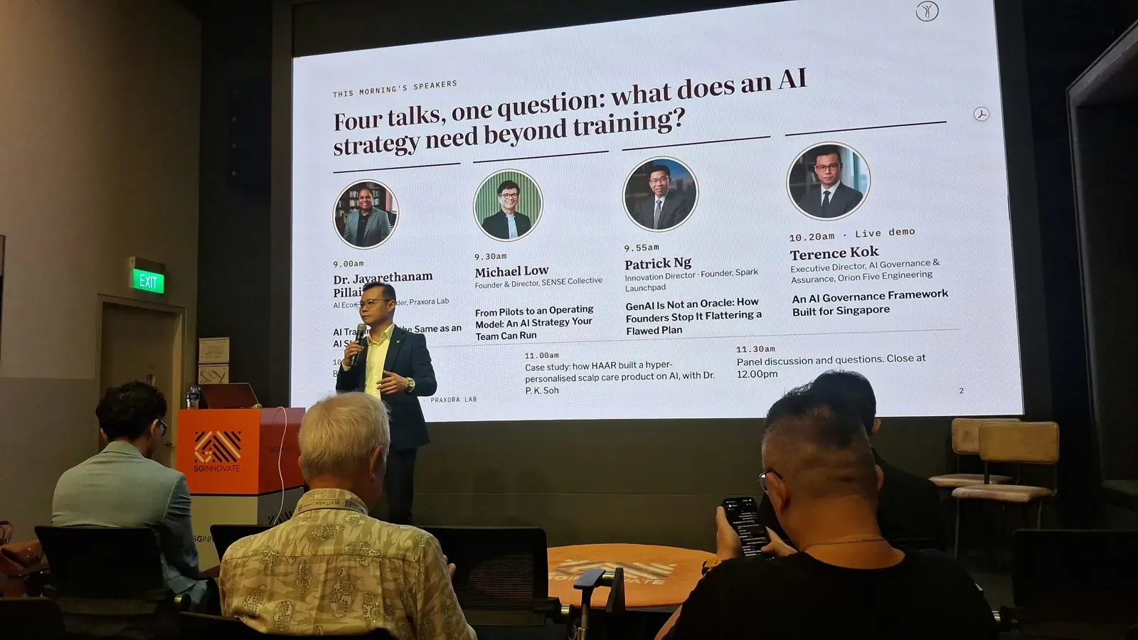 A speaker at the lectern beside a screen showing the morning's four talks and their speakers, with attendees seated in the foreground.