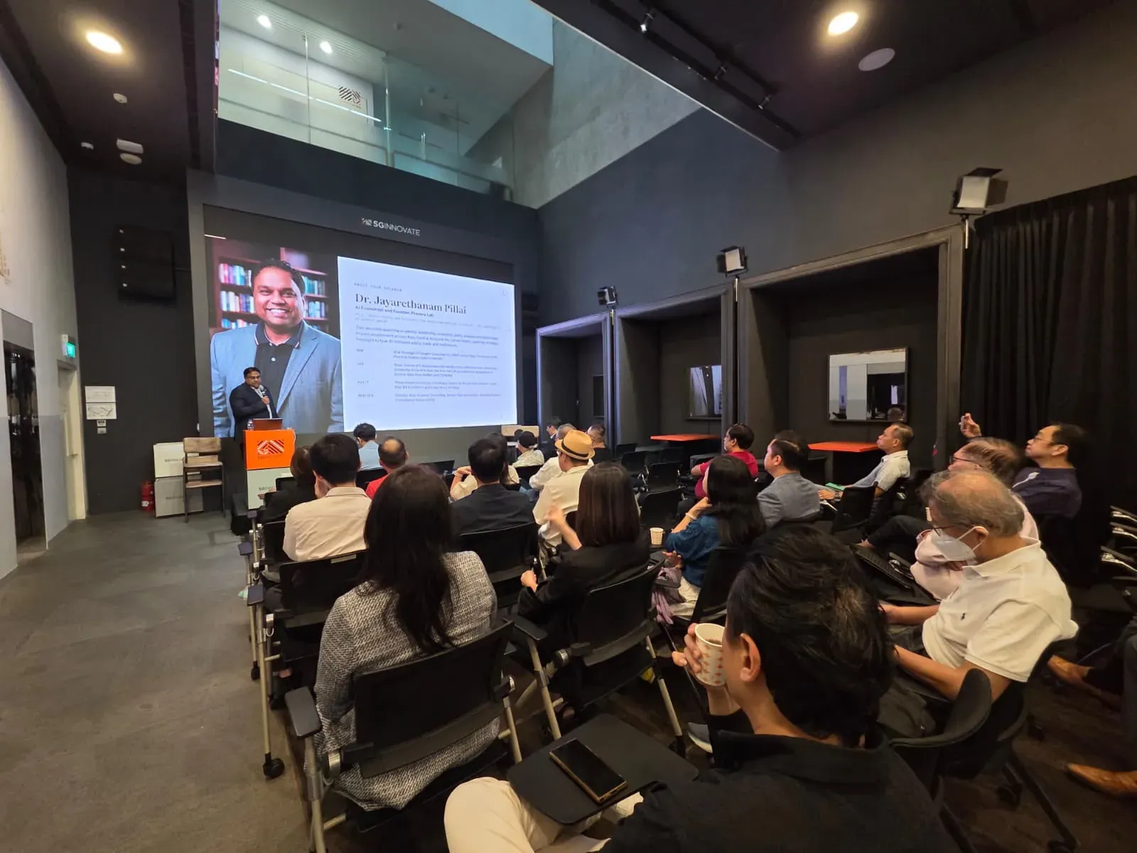 A full room at SGInnovate seen from the back, rows of attendees facing a large screen where Dr. Jayarethanam Pillai's speaker profile is displayed as he opens the morning from the lectern.